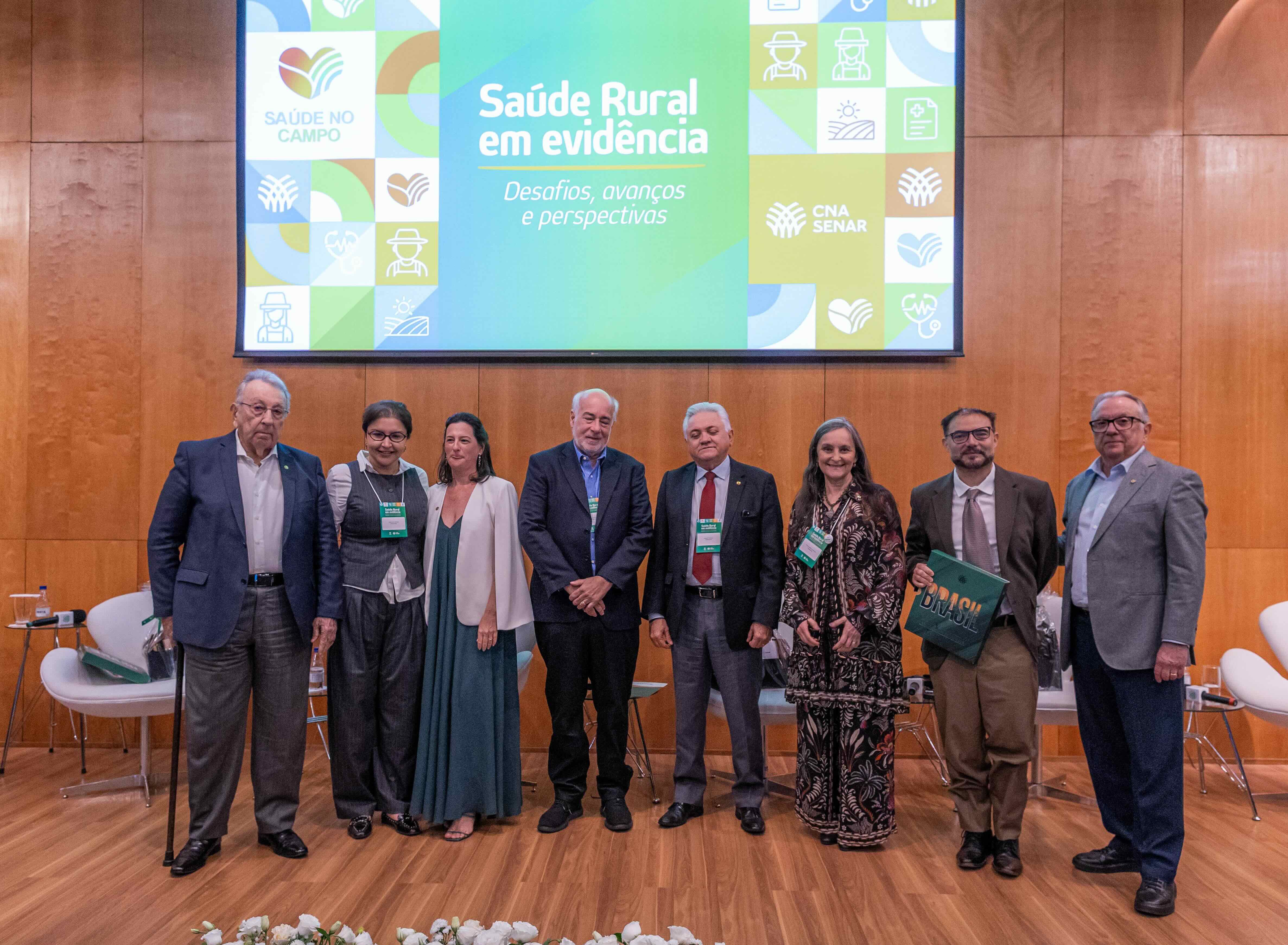Oito palestrantes e organizadores em pé no palco diante do telão com os dizeres Saúde Rural em Evidência.